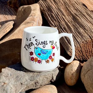 May include: White ceramic mug featuring a blue bird singing with musical notes above it. The mug is inscribed with the words "Then Sings my Soul".