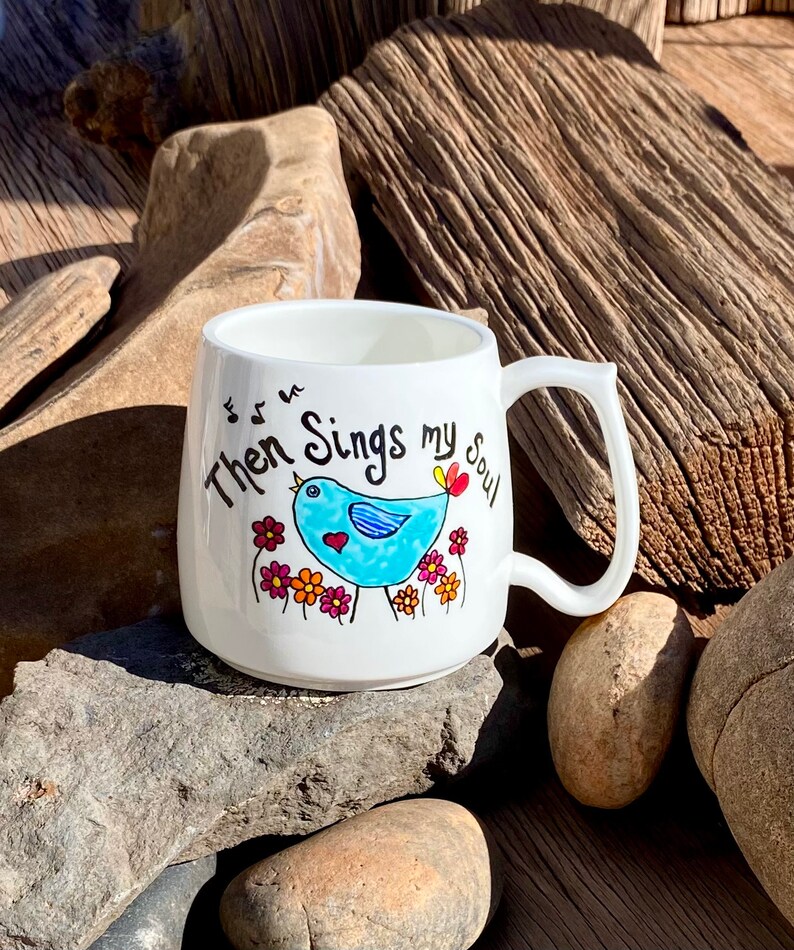 May include: White ceramic mug featuring a blue bird singing with musical notes above it. The mug is inscribed with the words "Then Sings my Soul".