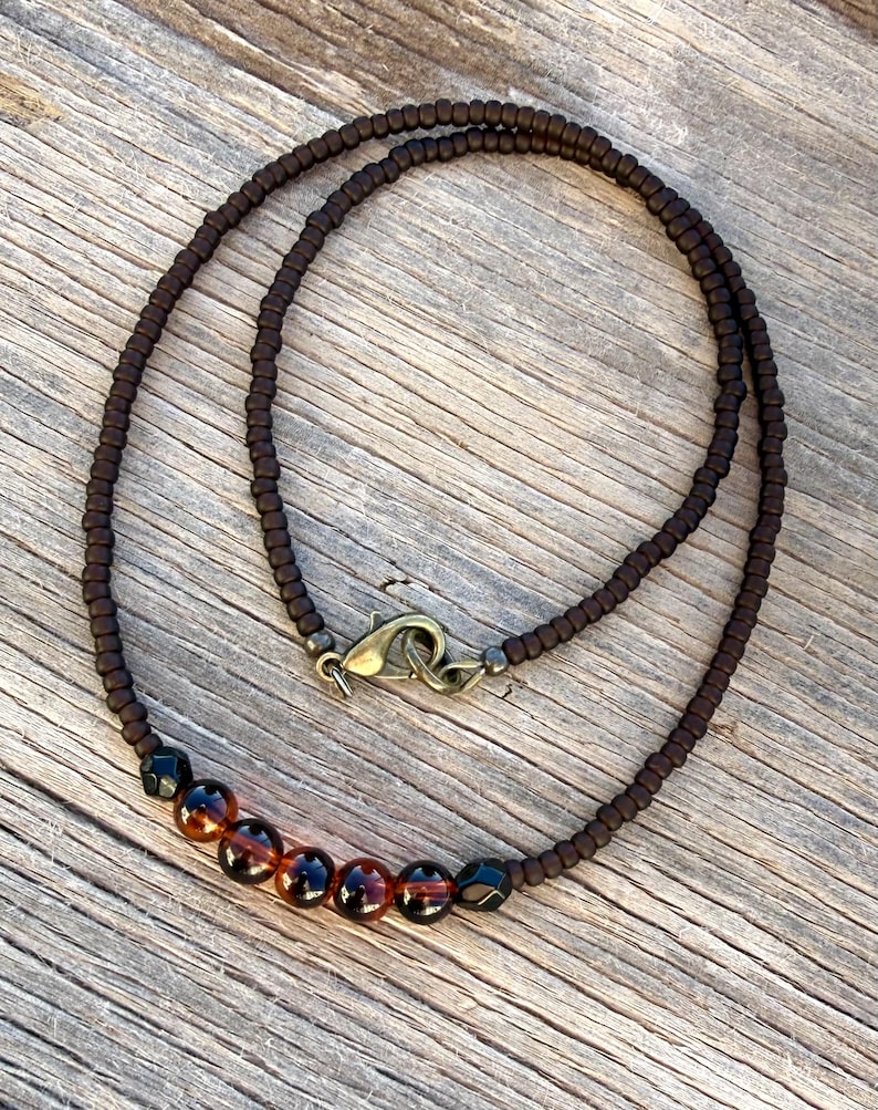 May include: A brown beaded necklace with a bronze clasp. The necklace features a row of amber and black beads. The necklace is displayed on a wooden surface.