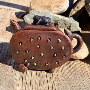 May include: A brown ceramic teapot with a frog on top. The teapot has a rounded body with a spout and handle. The teapot is decorated with small, round holes and gold-colored dots.