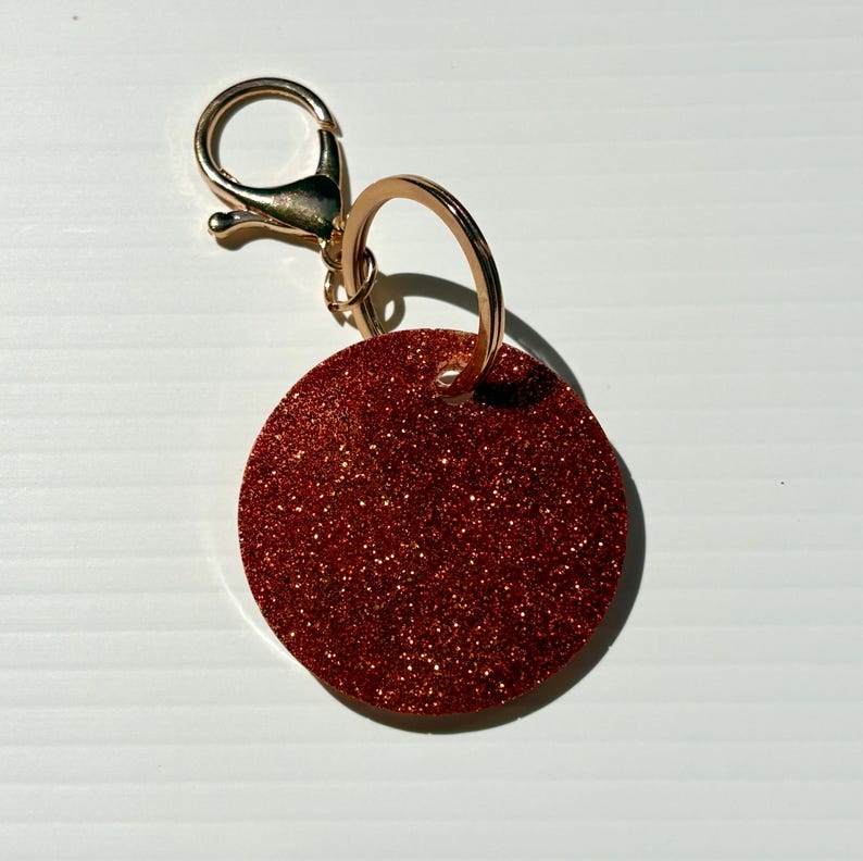 May include: A round, red glitter keychain tag with a gold-colored clasp and keyring. The tag is a solid circle shape and sparkles with glitter. The clasp is a lobster claw style.