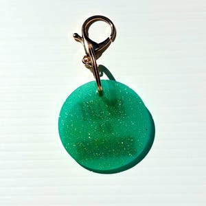 May include: A round, green keychain charm with gold glitter and a gold-colored clasp. The charm is a solid, opaque green with flecks of gold glitter throughout. The clasp is attached to the charm with a small gold ring.