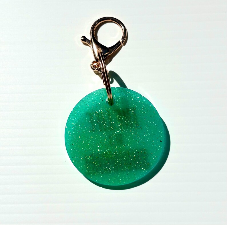 May include: A round, green keychain charm with gold glitter and a gold-colored clasp. The charm is a solid, opaque green with flecks of gold glitter throughout. The clasp is attached to the charm with a small gold ring.