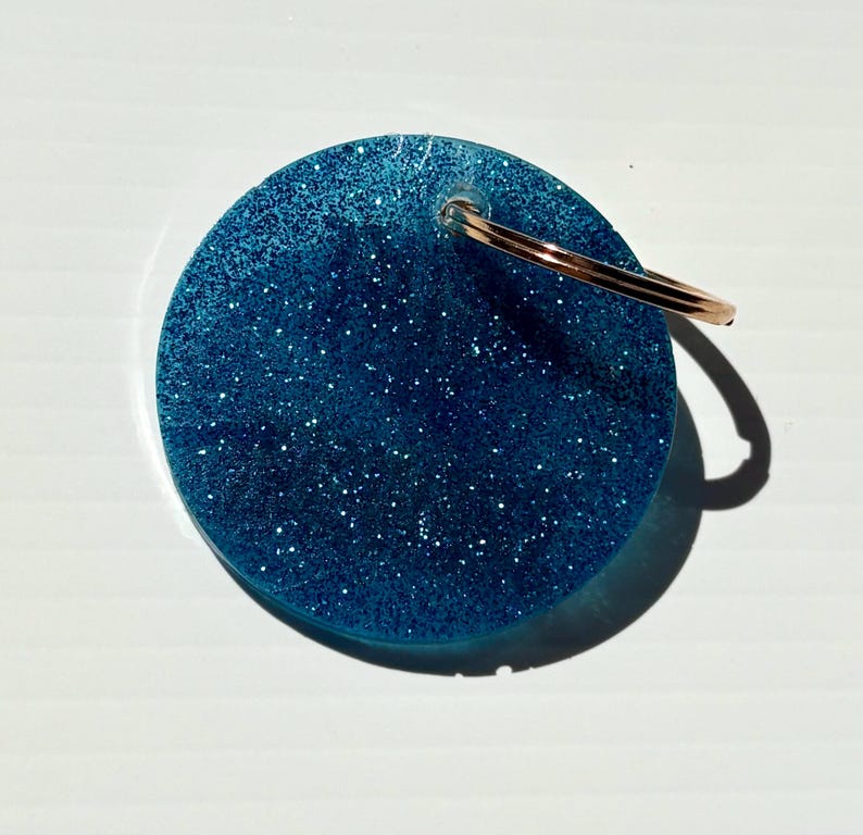 May include: A round, blue glitter keychain with a gold-colored keyring. The keychain is a solid circle with a sparkly, glittery texture. The keyring is attached at the top of the circle.