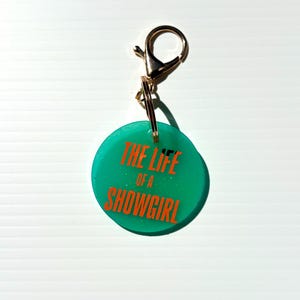 May include: A teal round keychain with the text "THE LIFE OF A SHOWGIRL" in orange. The keychain has a gold-colored clasp and ring.