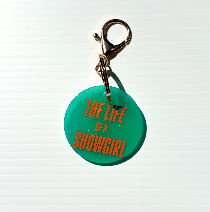 May include: A teal round keychain with the text "THE LIFE OF A SHOWGIRL" in orange. The keychain has a gold-colored clasp and ring.