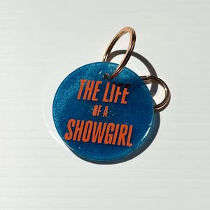 May include: A round, blue keychain with the text "THE LIFE of a SHOWGIRL" in orange lettering. The keychain has a gold-colored ring and a smaller ring attached.