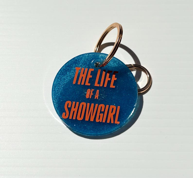 May include: A round, blue keychain with the text "THE LIFE of a SHOWGIRL" in orange lettering. The keychain has a gold-colored ring and a smaller ring attached.