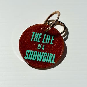 May include: A round, glittery, reddish-brown keychain with a gold-colored ring. The keychain features the text "THE LIFE of a SHOWGIRL" in a bright teal color. The background is a solid white.