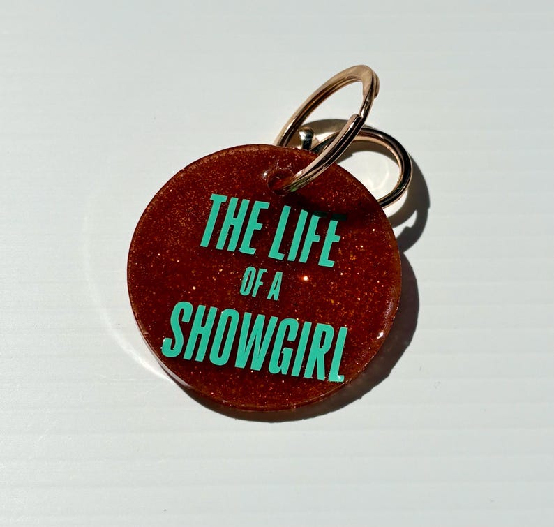 May include: A round, glittery, reddish-brown keychain with a gold-colored ring. The keychain features the text "THE LIFE of a SHOWGIRL" in a bright teal color. The background is a solid white.