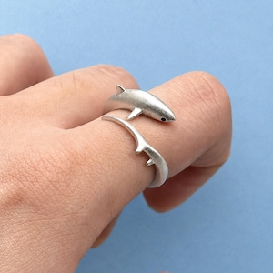 May include: A silver-toned wrap ring featuring a shark design. The ring is shaped like a shark with its body curving around the finger, showcasing a dorsal fin and a tail fin. The metal has a matte finish, and the shark has a small black eye detail.