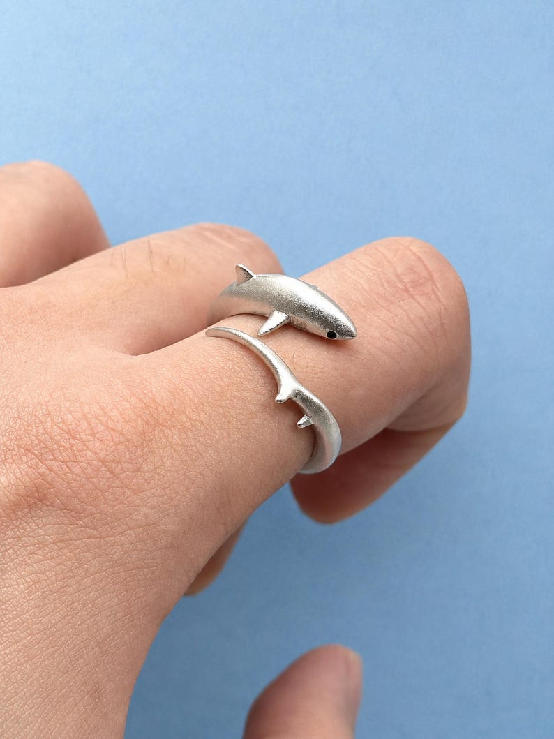 May include: A silver-toned wrap ring featuring a shark design. The ring is shaped like a shark with its body curving around the finger, showcasing a dorsal fin and a tail fin. The metal has a matte finish, and the shark has a small black eye detail.