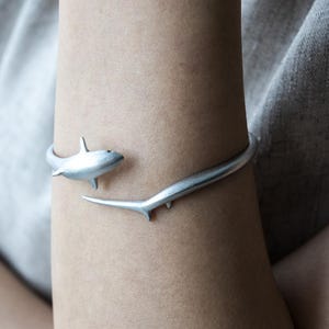 Op de afbeelding: Een zilverkleurige metalen armband met een haai-ontwerp. De armband is open, waarbij het ene uiteinde de vorm heeft van een haai en het andere uiteinde uitloopt in een dunne, gebogen, staartachtige verlenging die om de pols heen valt.