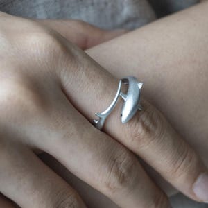 May include: A silver-toned open ring featuring a shark design. The ring is shaped like a shark with its body curving around the finger, with the tail end extending to form the other side of the band. The metal has a matte, brushed finish.