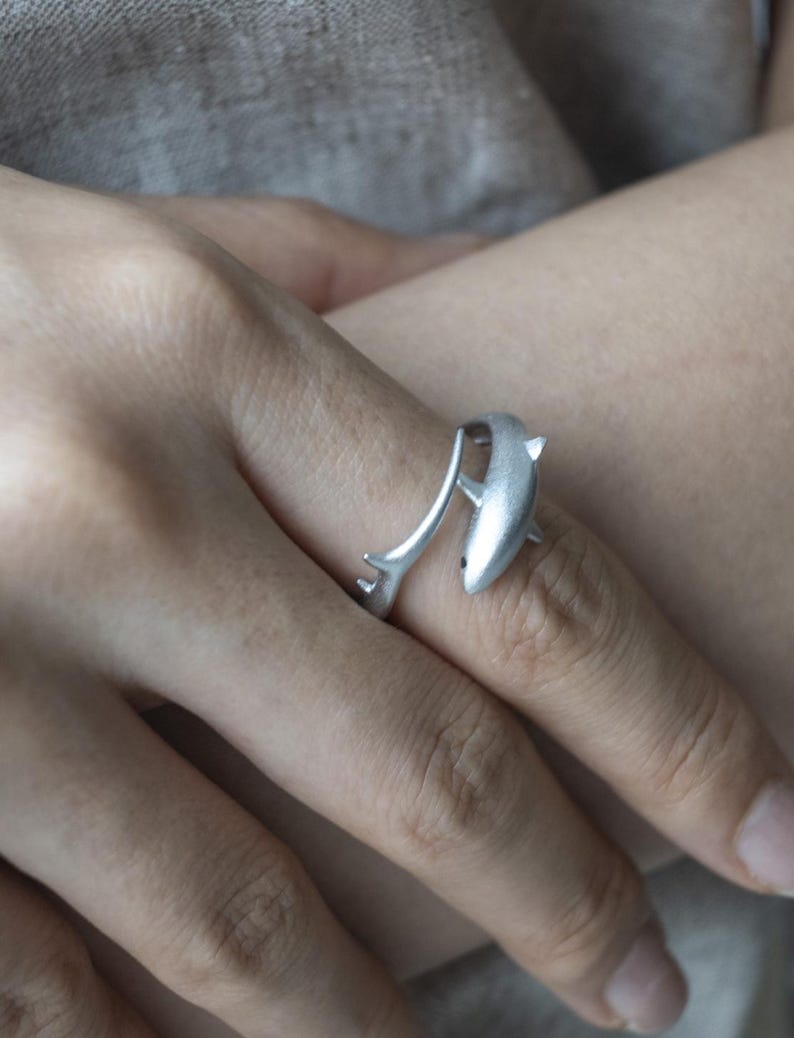 May include: A silver-toned open ring featuring a shark design. The ring is shaped like a shark with its body curving around the finger, with the tail end extending to form the other side of the band. The metal has a matte, brushed finish.