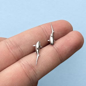 May include: A pair of silver-toned stud earrings shaped like sharks. Each earring features a textured, matte finish with a pointed dorsal fin, side fins, and a long, slender tail. The shark studs are oriented in opposite directions, showcasing a detailed aquatic design.