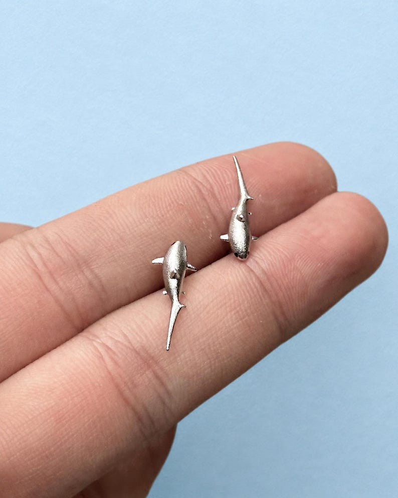 May include: A pair of silver-toned stud earrings shaped like sharks. Each earring features a textured, matte finish with a pointed dorsal fin, side fins, and a long, slender tail. The shark studs are oriented in opposite directions, showcasing a detailed aquatic design.