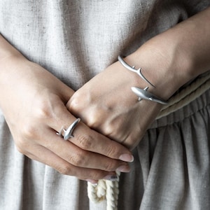 May include: A matte silver-toned open cuff bracelet and a matching ring, both featuring a shark-shaped design. The jewelry pieces have a smooth, metallic finish and are worn on the wrist and finger, showcasing a sleek, minimalist aesthetic.
