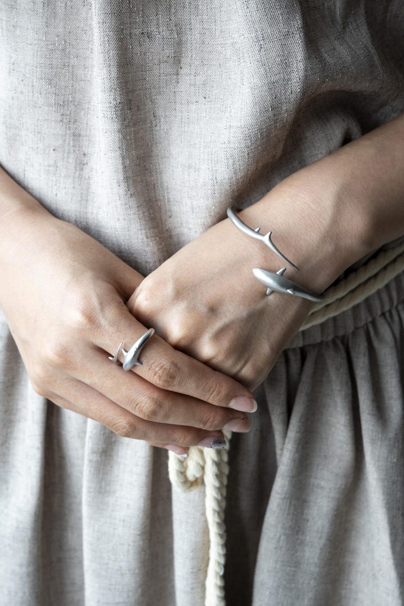May include: A matte silver-toned open cuff bracelet and a matching ring, both featuring a shark-shaped design. The jewelry pieces have a smooth, metallic finish and are worn on the wrist and finger, showcasing a sleek, minimalist aesthetic.
