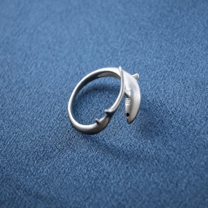 May include: A silver-toned metal ring featuring a wrap-around design shaped like a shark. The shark's head is positioned at one end of the band, while the tail fin is at the other, creating an open-ended silhouette with a smooth, matte finish.