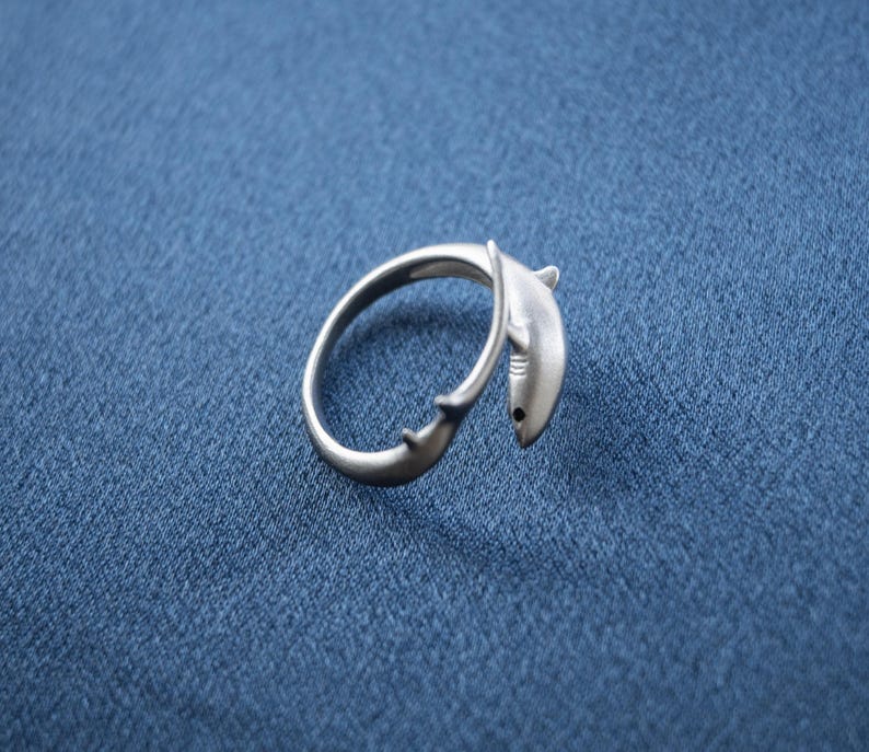 May include: A silver-toned metal ring featuring a wrap-around design shaped like a shark. The shark's head is positioned at one end of the band, while the tail fin is at the other, creating an open-ended silhouette with a smooth, matte finish.