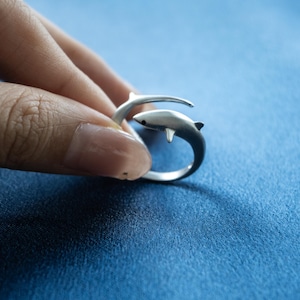 May include: A silver-toned open ring featuring a shark design that wraps around the finger. The shark has a smooth, matte finish with a small black dot for an eye and a dorsal fin. The band is adjustable, allowing the two ends to overlap slightly.
