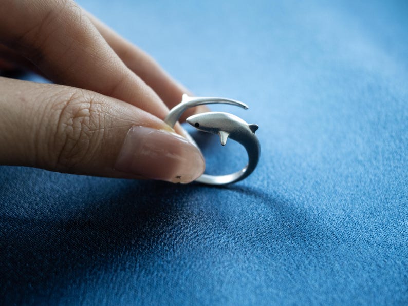 May include: A silver-toned open ring featuring a shark design that wraps around the finger. The shark has a smooth, matte finish with a small black dot for an eye and a dorsal fin. The band is adjustable, allowing the two ends to overlap slightly.