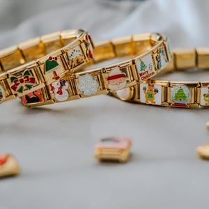 May include: Gold-toned charm bracelets with Christmas-themed charms. Charms include a snowman, Santa Claus, Christmas tree, and gift boxes. The bracelets and charms are on a light gray surface.