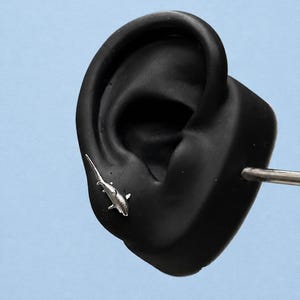 May include: A silver-toned shark-shaped stud earring positioned on a black ear-shaped display model. The shark design features a pointed dorsal fin, pectoral fins, and a long tail, with a metallic finish that reflects light against the dark surface.