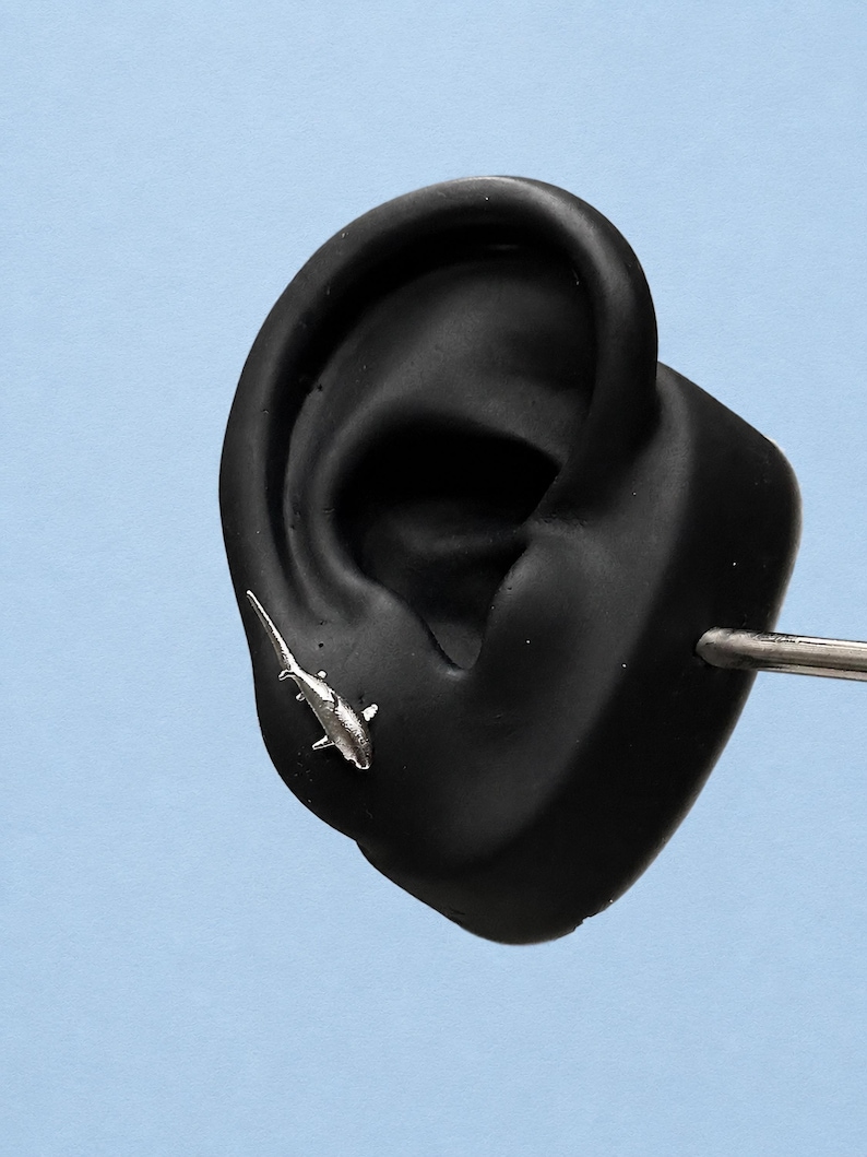 May include: A silver-toned shark-shaped stud earring positioned on a black ear-shaped display model. The shark design features a pointed dorsal fin, pectoral fins, and a long tail, with a metallic finish that reflects light against the dark surface.