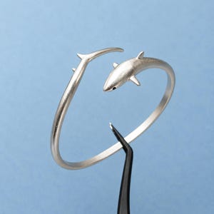 May include: A silver-toned open cuff bracelet featuring a shark design. The bracelet has a matte finish and a curved band that tapers into a shark head on one end and a pointed tail fin on the other. The shark has a small black eye detail.