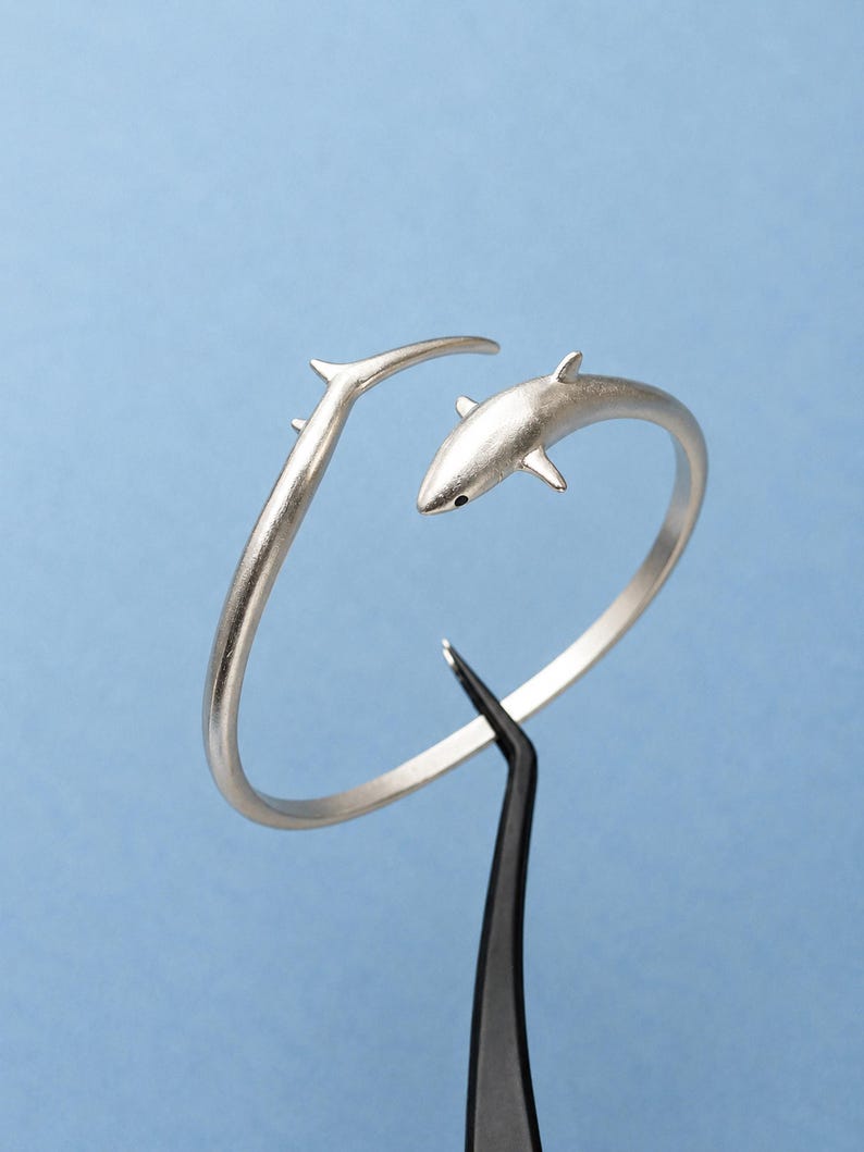 May include: A silver-toned open cuff bracelet featuring a shark design. The bracelet has a matte finish and a curved band that tapers into a shark head on one end and a pointed tail fin on the other. The shark has a small black eye detail.