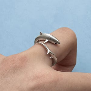 May include: A silver-toned wrap ring featuring a shark design. The shark's body forms the band of the ring, with the head and tail meeting at the front. The metal has a matte finish, and the shark has a small, dark, circular eye detail.