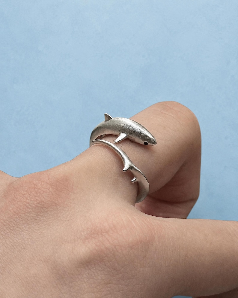 May include: A silver-toned wrap ring featuring a shark design. The shark's body forms the band of the ring, with the head and tail meeting at the front. The metal has a matte finish, and the shark has a small, dark, circular eye detail.