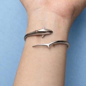 May include: A silver-toned metal cuff bracelet featuring a shark design. The bracelet is open-ended, with the head of the shark on one side and the tail fin on the other, wrapping around the wrist in a continuous, sleek, and polished metallic finish.
