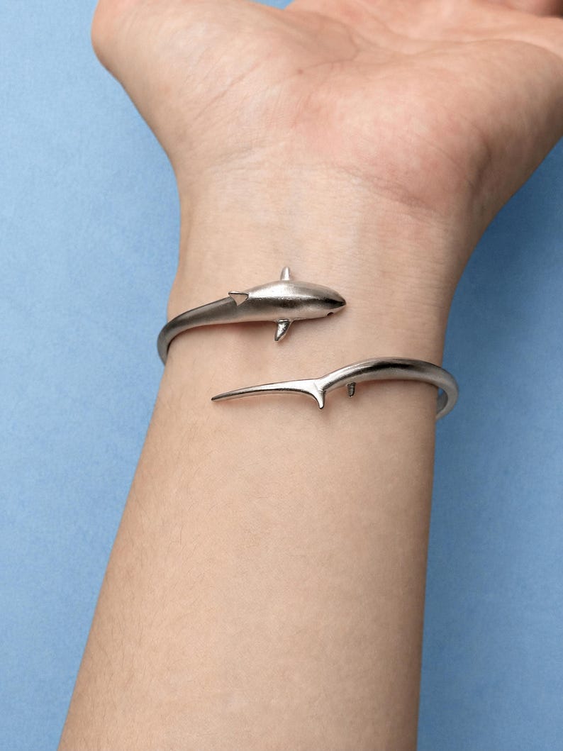 May include: A silver-toned metal cuff bracelet featuring a shark design. The bracelet is open-ended, with the head of the shark on one side and the tail fin on the other, wrapping around the wrist in a continuous, sleek, and polished metallic finish.