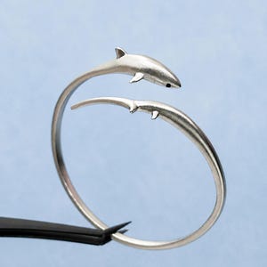 May include: A silver-toned open cuff bracelet featuring two shark figures facing each other. The shark at the top has a visible black eye detail, while the lower shark is depicted with a sleek, elongated body. The metal has a smooth, matte finish.