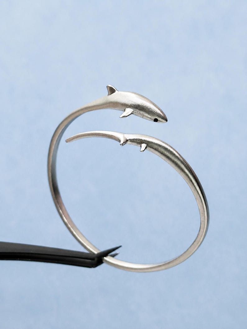 May include: A silver-toned open cuff bracelet featuring two shark figures facing each other. The shark at the top has a visible black eye detail, while the lower shark is depicted with a sleek, elongated body. The metal has a smooth, matte finish.