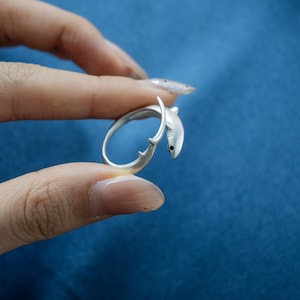 May include: A silver-toned wrap ring shaped like a shark, featuring a smooth metallic finish, a curved body that forms the band, and a small black dot detail representing the eye on the shark's head, which rests against the tail end.
