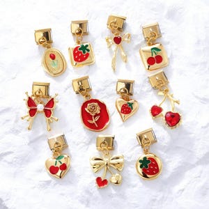 Puede incluir: Una colección de dijes dorados con varios diseños, incluyendo fresas, cerezas, rosas, mariposas y lazos. Cada dije tiene un cierre rectangular en la parte superior. Los dijes son de color rojo, dorado y blanco.
