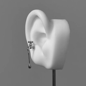 May include: A silver-toned earring shaped like a small water faucet attached to the earlobe. A delicate silver chain hangs from the spout of the faucet, ending in a small, polished silver drop-shaped charm that mimics a single falling water droplet.