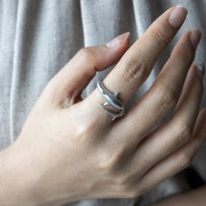 May include: A silver-toned wrap ring featuring a shark design that encircles the finger. The shark's body forms the band, with the head and tail overlapping to create an open-ended, adjustable style with a smooth, matte metallic finish.