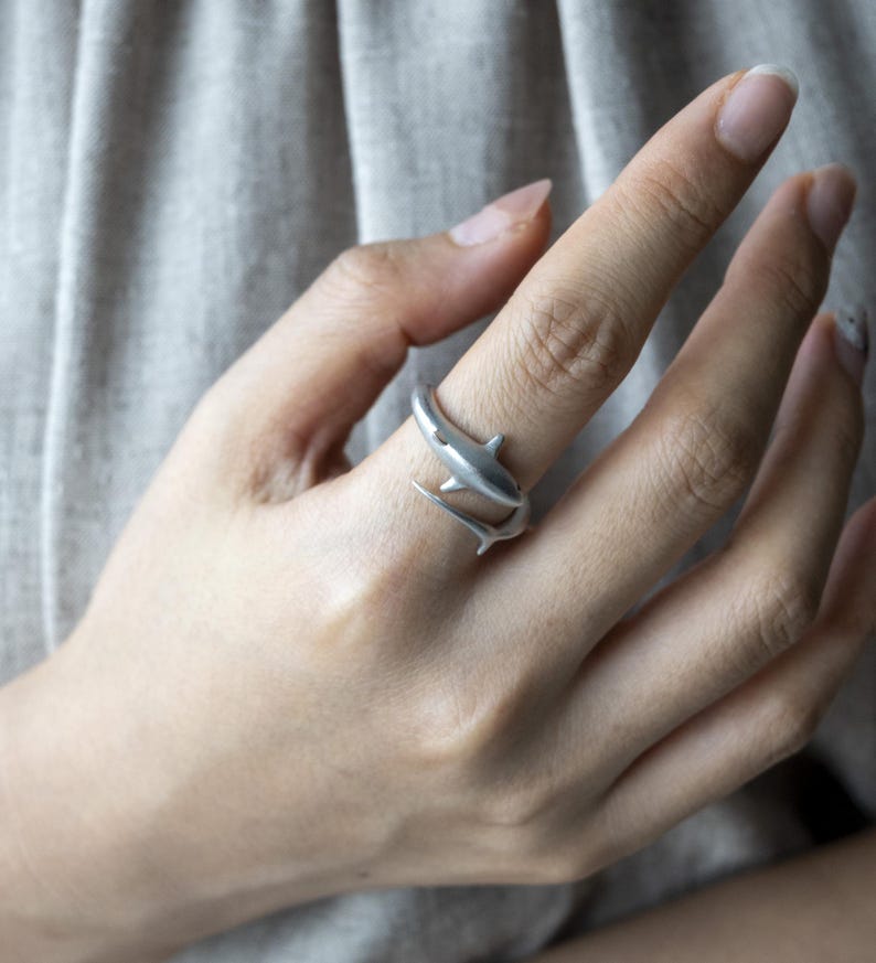May include: A silver-toned wrap ring featuring a shark design that encircles the finger. The shark's body forms the band, with the head and tail overlapping to create an open-ended, adjustable style with a smooth, matte metallic finish.