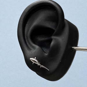 May include: A silver-toned stud earring shaped like a hammerhead shark. The earring features a textured metallic finish and is positioned on a black ear-shaped display model. The shark design includes a wide head, dorsal fin, and a long tail.