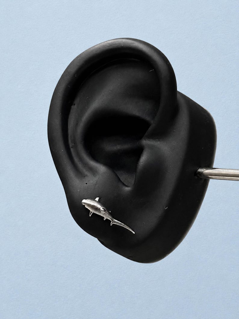 May include: A silver-toned stud earring shaped like a hammerhead shark. The earring features a textured metallic finish and is positioned on a black ear-shaped display model. The shark design includes a wide head, dorsal fin, and a long tail.