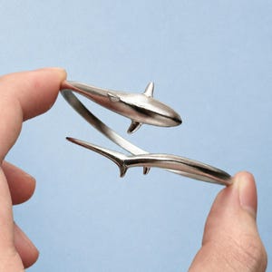 May include: A silver-toned metal wrap bracelet featuring a shark design. The bracelet is shaped like a shark with the head and dorsal fin on one end and the tail section on the other, creating an open-ended, adjustable circular band structure.