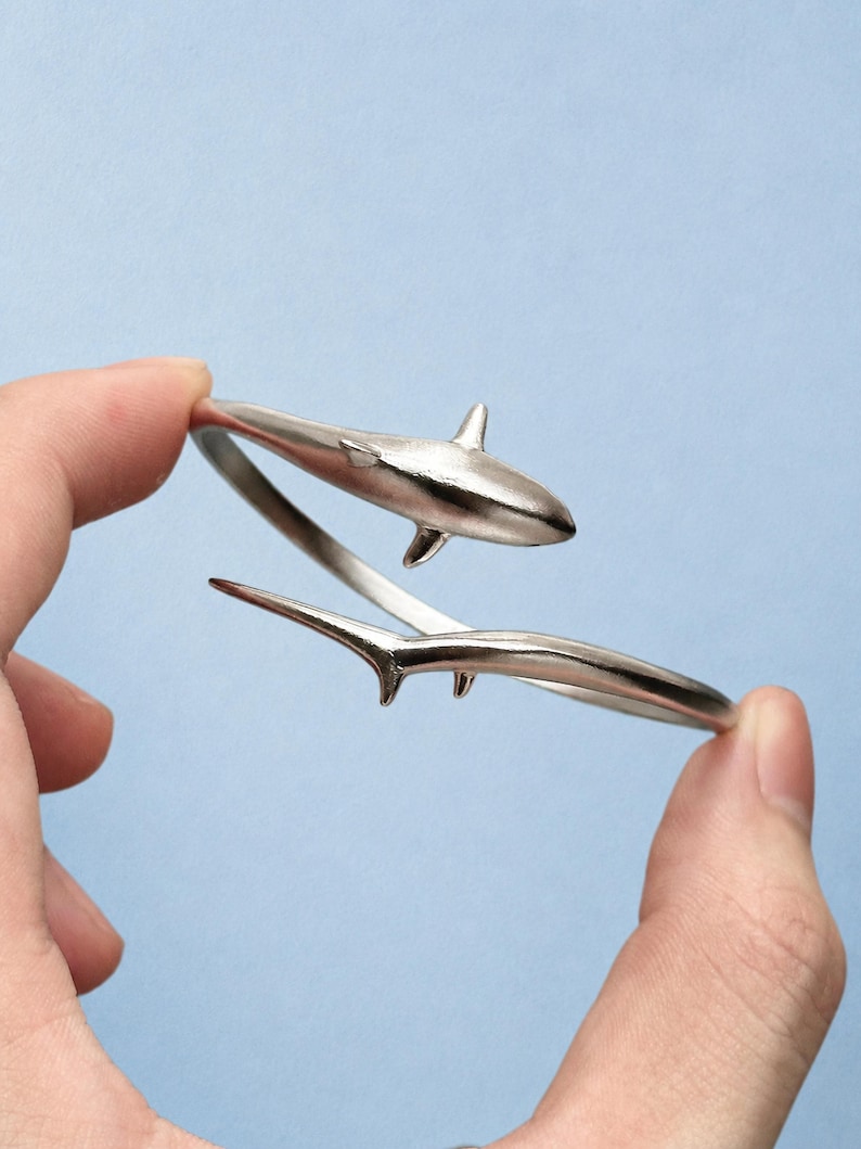 May include: A silver-toned metal wrap bracelet featuring a shark design. The bracelet is shaped like a shark with the head and dorsal fin on one end and the tail section on the other, creating an open-ended, adjustable circular band structure.