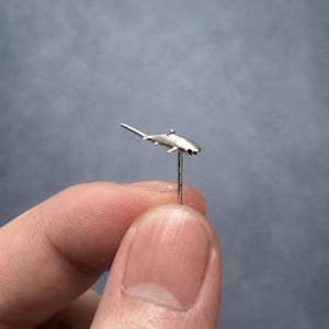 May include: A small silver-toned shark-shaped stud earring with a thin metal post extending downwards from the center of the shark's body. The shark figure is metallic and textured, featuring a pointed tail, dorsal fin, and pectoral fins.