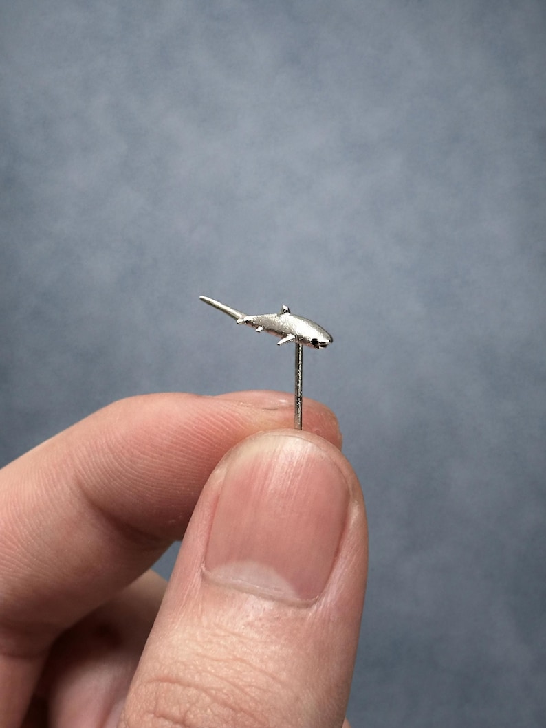 May include: A small silver-toned shark-shaped stud earring with a thin metal post extending downwards from the center of the shark's body. The shark figure is metallic and textured, featuring a pointed tail, dorsal fin, and pectoral fins.
