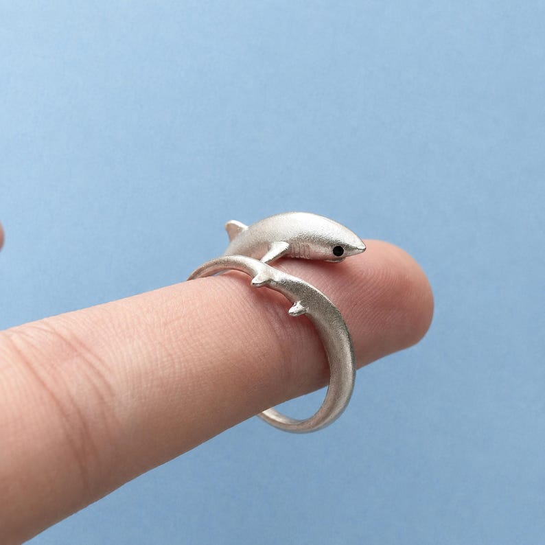 May include: A silver-toned wrap ring featuring a shark design. The ring is shaped like a shark with its body forming the band and its head and tail meeting at the front. The metal has a matte, brushed finish and a small black dot for the eye.